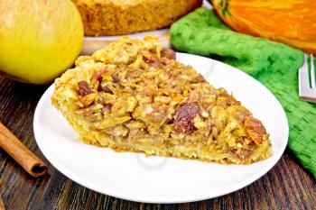Piece of pie with apples, pumpkin, raisins and nuts in a white plate, cinnamon, napkin on a background of a dark wooden board