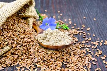 Flaxseed flour in a spoon, seeds in a bag and on a table, blue linen flower on a background of a wooden board