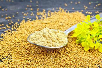 Mustard powder in a metal spoon, seeds and mustard flower on a wooden board background