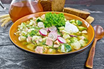 Cold soup okroshka from sausage, potato, egg, radish, cucumber, greens and kvass in a bowl, bread and jug with drink on background of dark wooden board