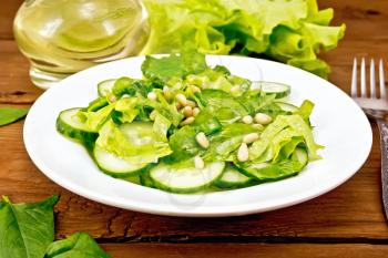 Salad from spinach, fresh cucumbers, rukkola salad, cedar nuts and spring onions, seasoned with vegetable oil on a plate, fork in the background of the board