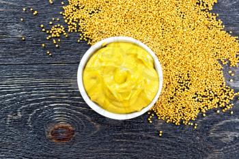 Mustard sauce in a white bowl and seeds on a wooden board background on top