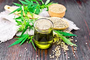 Hemp oil in a glass jar, grain in a spoon and flour in a bowl on sackcloth, cannabis leaves and stalks on a wooden board background