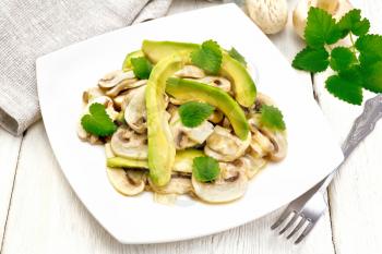 Salad of avocado and raw champignons, seasoned with lemon juice and vegetable oil with mint leaves, towel and a fork on the background of light wooden board
