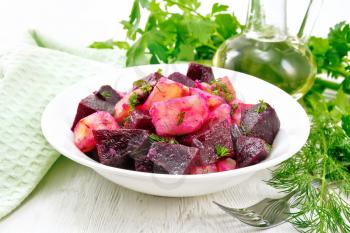 Salad of beets and potatoes, seasoned with vegetable oil and vinegar in a plate, napkin, parsley and dill on the background of a light wooden board