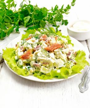 Salad from salmon, cucumber, eggs and avocado, dressed with mayonnaise on lettuce leaves in a plate, napkin, dill, parsley and fork on a white wooden board background