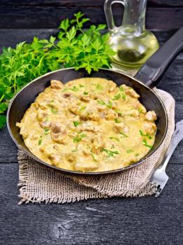 Meat stewed with cream in an old frying pan on burlap, parsley, fork and vegetable oil on a wooden board background