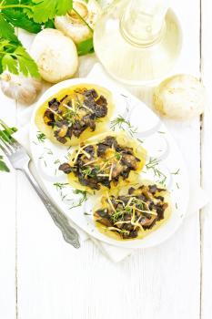 Potatoes stuffed with mushrooms, fried onions and cheese in a plate on napkin, vegetable oil in decanter, parsley, garlic and a fork on wooden board background from above