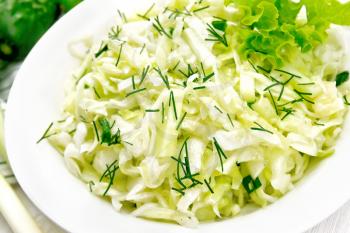 Salad of fresh cabbage, green onions and cucumber with vinegar and vegetable oil dressing in a plate, towel, dill, lettuce and fork on wooden board background