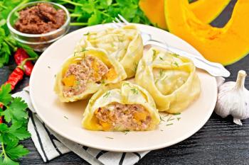 Manty steamed pies with minced meat and pumpkin in a plate on a kitchen towel, spicy sauce, parsley, garlic and pepper on dark wooden board background