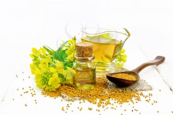 Mustard oil in a glass bottle and gravy boat, grains in a spoon and on a burlap napkin, yellow mustard flowers on wooden board background