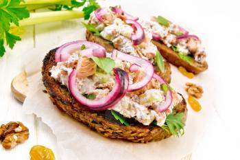 Salmon, petiole celery, raisins, walnuts, red onions and curd cheese salad on toasted bread with green lettuce leaves on parchment on a light wooden board background