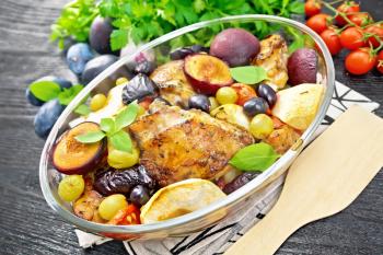 Baked chicken with tomatoes, apples, plums and grapes in a glass roaster on a towel, garlic, parsley and basil on background of dark wooden board