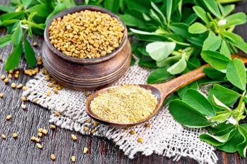 Ground fenugreek in a spoon and seeds in a bowl on a burlap napkin with leaves on wooden board background