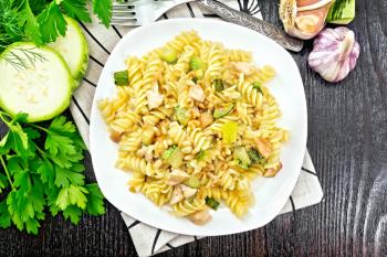 Fusilli pasta with chicken breast, zucchini, cream and pine nuts in a plate on a napkin, garlic, fork and parsley on a wooden board background from above