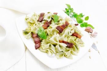 Farfalle pasta with pesto sauce, fried bacon and basil in a plate, garlic, fork and napkin on wooden board background