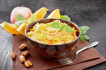 Pumpkin, carrot and apple salad with pecans seasoned with vegetable oil in a bowl on a napkin, mint on dark wooden board background