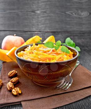 Pumpkin, carrot and apple salad with pecans seasoned with vegetable oil in a bowl on a towel, mint on black wooden board background