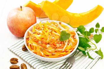 Pumpkin, carrot and apple salad with pecans seasoned with vegetable oil in a bowl on a towel, mint on wooden board background