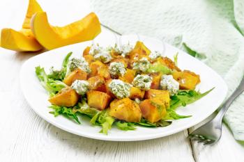 Salad of baked pumpkin, arugula with balls of salt cheese, seasoned with honey, mustard, garlic and vegetable oil in a plate, napkin and fork on light wooden board background