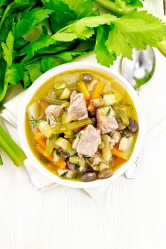 Eintopf soup of pork, celery, beans, carrots and potatoes with leek in a white bowl on a napkin on the background of light wooden board from above
