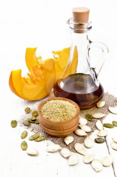 Pumpkin flour in a bowl and oil in a decanter on burlap, seeds on table and slices of vegetable on wooden board background