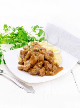 Beef goulash in tomato sauce with mashed potatoes in a plate, napkin, parsley and fork on background of light wooden board