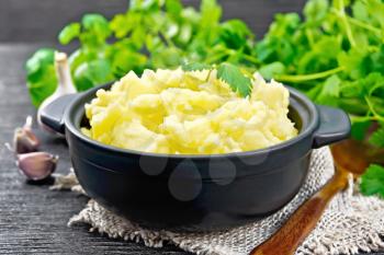 Mashed potatoes in a black saucepan and a spoon on burlap, garlic, parsley on wooden board background