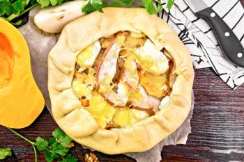 Pie of pumpkin, pear, soft cheese and walnuts on parchment, towel and knife, parsley on a wooden board background from above