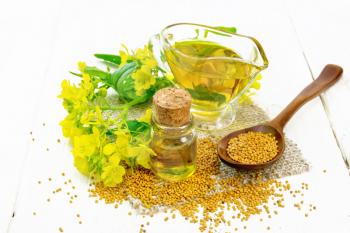 Mustard oil in a glass bottle and gravy boat, grains in a spoon and burlap, yellow mustard flowers on white wooden board background