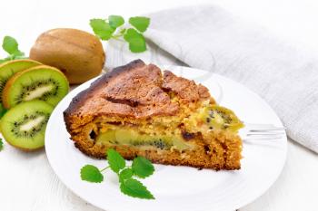 Piece of sweet cake with kiwi and honey, mint and a fork in a plate, a towel on a background of a light wooden board
