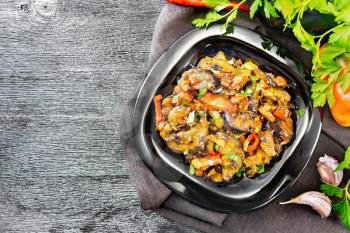 Vegetable ragout with eggplant, tomatoes, sweet and hot peppers, onions, carrots, fried with herbs and spices in a plate on a towel, garlic, parsley on wooden board background from above