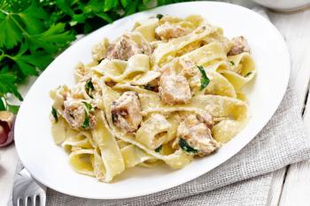 Tagliatelle pasta with salmon, cream, garlic and herbs in a plate on a napkin, fork, parsley and basil on wooden board background