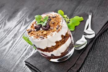 Dessert Black Forest of cherries, chocolate biscuit and soft cottage cheese with cream in a glass on napkin, mint, berries in a bowl and a spoon on dark wooden board background