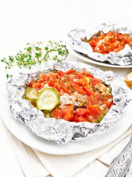 Pink salmon with zucchini, tomatoes, onions, garlic and thyme, baked in foil on a plate, towel, fork and bread on a wooden board background
