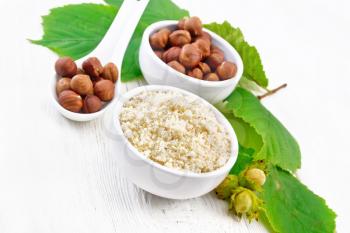 Flour and hazelnuts in two bowls, a spoon with peeled walnut kernels and a branch of filbert with green leaves on wooden board background