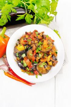 Vegetable ragout with eggplant, tomatoes, sweet and hot peppers, onions, carrots, fried with herbs and spices in plate on a towel, garlic, parsley on white wooden board background from above