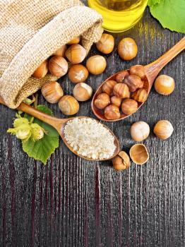 Hazelnut flour in a spoon, nuts in a bag, spoon and on the table, oil in glass jar and filbert branch with green leaves on a wooden board background from above