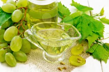 Grape oil in a gravy boat and a jar on sacking, berries of green grapes on the background of light wooden board