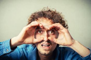 Curly young man makes the binoculars around your eyes from the hands. On a gray background