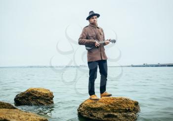 young man playing ukulele on stones at the lake