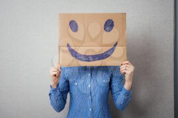 Young woman holding a cardboard with a smiley face on it in front of her head.