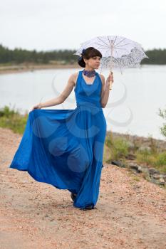 Young slim girl in a blue dress with an umbrella walking on a country road.