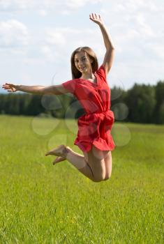 Young girl in a red dress jumping in a field with coniferous forest.