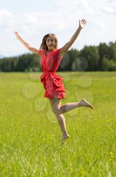 Young girl in a red dress jumping in a field with coniferous forest