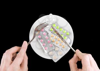Coloured pills in white plate with fork and spoon in hands, on black background.