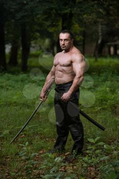 Action Hero Muscled Man Holding A Ancient Sword - Standing In Forest Wearing Leather Pants
