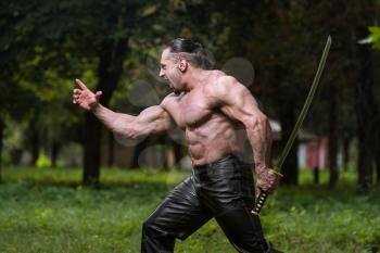 Action Hero Muscled Man Holding A Ancient Sword - Standing In Forest Wearing Leather Pants