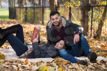 Beautiful Young Couple Sitting In The Park On A Beautiful Autumn Day - They Are Reading A Book