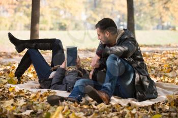 Beautiful Young Couple Sitting In The Park On A Beautiful Autumn Day - They Are Using Internet Via Digital Tablet